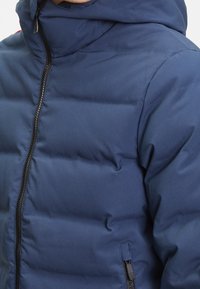 Tmavě modrá prošívaná bunda s vysokým límcem, která má přední zip, prošívanou strukturu a boční kapsu na zip. Hladký, voděodolný materiál.