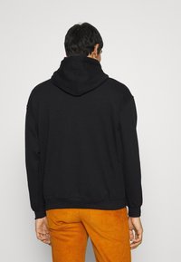 Sudadera negra con capucha de corte relajado, puños y dobladillo acanalados. Llevada con pantalones de pana naranja, vista desde atrás.