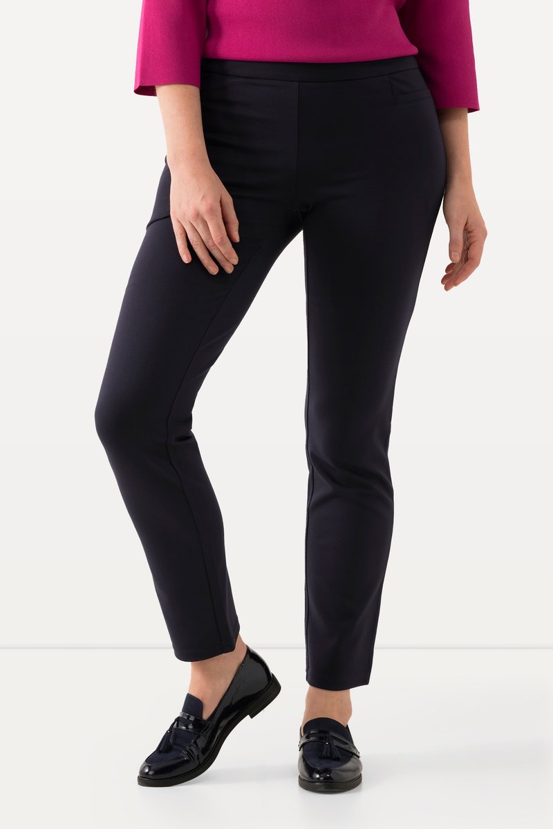 Femme portant un pantalon noir slim, des mocassins noirs à pampilles et un top rose à manches longues, debout, jambes croisées.