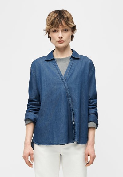 Jeune personne aux cheveux courts portant une chemise en denim ample sur un haut gris et un pantalon blanc, debout devant un fond blanc uni.