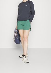Sudadera de color azul oscuro, pantalones cortos deportivos verdes, zapatillas deportivas de color claro. Mochila con estampado floral, de pie sobre un fondo neutral.