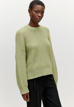 Lichtgroene gebreide trui met een ronde hals, geribbelde mouwen en zoom. Draagt met zwarte broek, toont een textuurdesign.