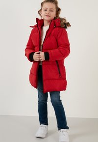 Rød polstret parka-jakke med høj krave, sorte lynlåse og ribstrikkede manchetter. Bæres over en lys sweater og parret med blå jeans og hvide sneakers.