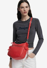 Borsa a spalla in pelle rossa con forma arrotondata, dotata di tasca con zip, logo in rilievo e tracolla regolabile con hardware argentato.