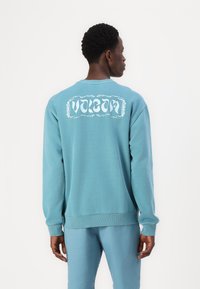 Modrá bavlnená mikina s dlhými rukávmi, rebrovanými manžetami a spodným lemom. Na zadnej strane sa nachádza veľké biele logo "Volcom" v ozdobnom ráme.
