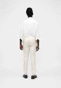 Άνδρας με σκούρο δέρμα και κοτσίδες στέκεται με την πλάτη προς τα πίσω, φορώντας λευκό πουκάμισο με μακριά μανίκια, κρεμ παντελόνια, λευκές κάλτσες και σκούρα παπούτσια.