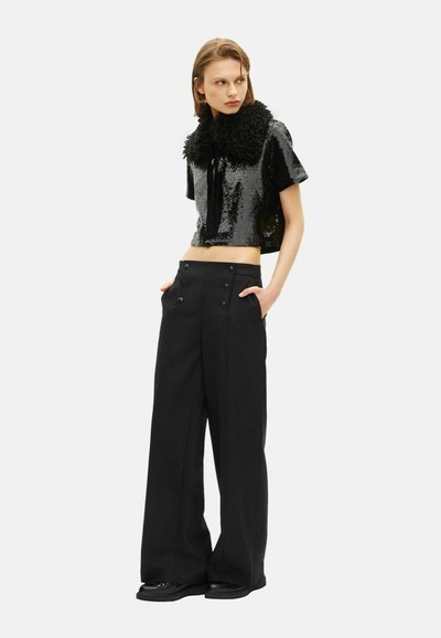 Donna che indossa pantaloni neri a gamba larga e un top corto nero con paillettes e un ampio colletto texturizzato, in piedi con le mani nelle tasche.