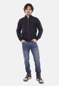 Giacca zip-up blu navy realizzata in tessuto morbido, con colletto alto, abbinata a jeans in denim azzurri e sneakers marroni con dettagli bianchi.