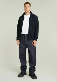 Giacca zip-up blu scuro sopra una t-shirt bianca, abbinata a jeans in denim scuro a taglio largo e sneakers nere chunky. Design semplice e casual.