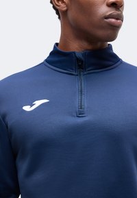 Gros plan d'une personne portant un haut de sport bleu marine à fermeture éclair quart, avec un logo blanc sur la poitrine gauche.