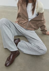 Braune Jacke mit Reißverschluss, weißes Tanktop, graue, weit geschnittene Jogginghose und braune Schnürschuhe. Lässiges Outfit mit weichen Texturen und entspannter Passform.