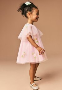 Rosa Tüllkleid mit floralen Stickereien, flatternden Ärmeln und einer taillierten Taille. Kombiniert mit silbernen Mary Jane Schuhen. Leichte und luftige Textur.