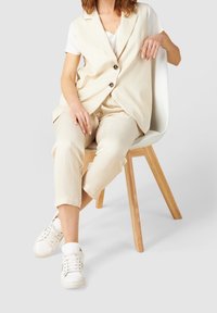 Béžová vesta a odpovídající kalhoty vyrobené z lehké tkaniny, doplněné o bílé tenisky. Židle má dřevěnou základnu a bílý sedák.