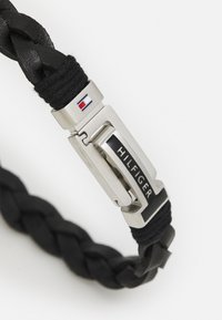 Svart flettet lærarmbånd med en sølvspenne som har svart og hvit aksent som viser "HILFIGER" og et lite logo i hjørnet.