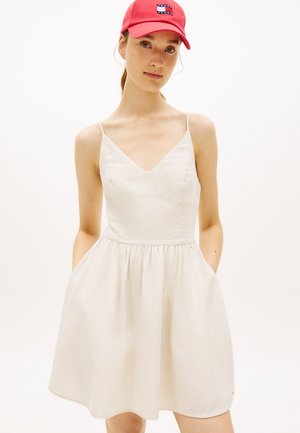 Junge Frau trägt ein hell gestreiftes, ärmelloses Kleid und eine rote Kappe mit Logo, Hände in den Taschen des Kleides, neutraler Hintergrund.