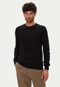 Schwarzer Strickpullover mit strukturiertem Zopfmuster, Rundhalsausschnitt und gerippten Bündchen. Getragen über beigen Hosen, mit einem kleinen Logodetail.