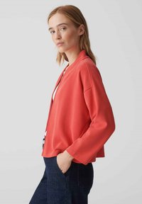 Veste bomber corail avec une coupe décontractée, présentant des épaules tombantes, des manches larges et une texture lisse. Portée par-dessus un haut blanc et un jean foncé.