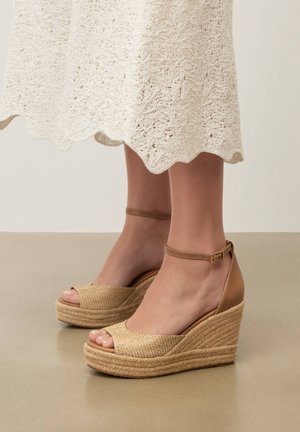 Picioare în sandale bej cu platformă împletită și bretele la gleznă, asortate cu tivul unei fuste albe croșetate, stând pe un podea netedă.