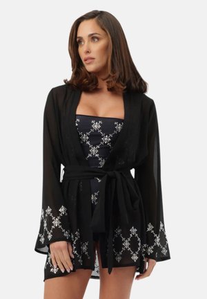 Donna che indossa un accappatoio nero trasparente con motivi geometrici argentati e vita annodata sopra un abito nero senza spalline abbinato.