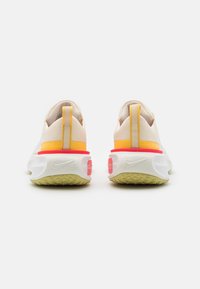 Des chaussures de course blanches avec un dessus texturé beige clair, des accents orange vif et rouge sur la semelle intermédiaire, et une semelle extérieure en caoutchouc verte.