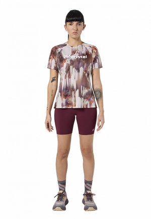 Mujer de pie de frente, vistiendo una camisa estampada abstracta multicolor, pantalones cortos de ciclismo granate, calcetines estampados y zapatillas deportivas.