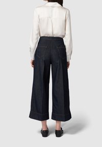 Tmavě modré džínové široké kalhoty s vysokým pasem, viditelnými zadními kapsami a kontrastním šitím. V páru se světlou košilí na knoflíky.