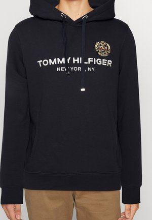 Sort Tommy Hilfiger-hoodie med hvid broderet logo og teksten New York, med et våbenskjoldsemblem på venstre bryst og forlommer.