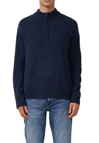 Mężczyzna ubrany w ciemnoniebieski sweter typu half-zip z ribbowanym kołnierzem, mankietami i dołem, w połączeniu z jasnoniebieskimi dżinsami, na białym tle.