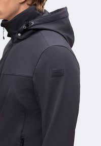 Homme portant une veste softshell noire à capuche avec fermeture éclair et écusson logo sur le haut de la manche, vu de profil.