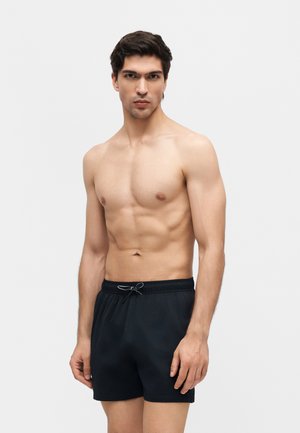 Muž bez trička s krátkymi tmavými vlasmi, oblečený v čiernych šortkách so šnúrkou, stojaci pred jednoduchým svetlým pozadím.