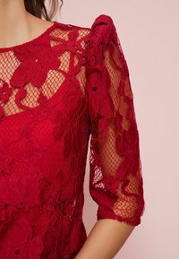 Haut en dentelle rouge avec des motifs floraux et des manches trois-quarts transparentes, doublé d'un tissu rouge uni et lisse en dessous.