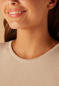 Rippentop in Beige mit einem runden Ausschnitt, der eine glatte Textur aufweist. Kleine goldene Steckerohrringe sind am rechten Ohr sichtbar.