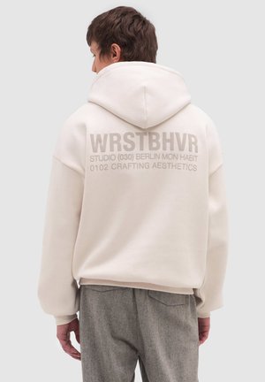Persona che indossa una felpa con cappuccio beige chiaro con cappuccio abbassato, con grande scritta WRSTBHVR e dettagli più piccoli su studio e luogo sul retro.