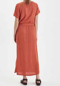 Maxi šaty v rzi oceánové barvě s krátkými rukávy, páskem v pase a jemným leskem. Mají volný střih a boční rozparky pro snadný pohyb.