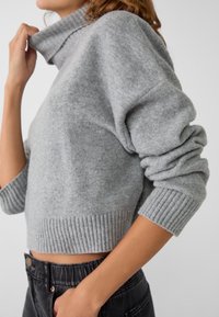 Pull en maille gris avec col montant et poignets côtelés. Le tissu semble doux, avec une coupe courte et une coupe décontractée.