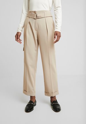 Person iført beige viddebukser med bæltestropper og manchetter, kombineret med sorte loafers og en hvid langærmet top.