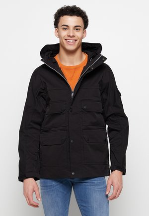 Black parka jacket with a hood, featuring four front pockets, a zip closure, and button accents. Worn over an orange shirt.