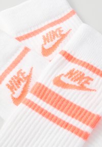 Baltos medvilninės kojinės su dviem horizontaliomis oranžinėmis juostelėmis ir oranžiniu "Nike" logotipu, išsiuvinėtu priekyje. Minkšta tekstūra ir raukšlėtas dizainas.