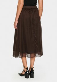 Bruine chiffon midi-rok met een elastische tailleband, met een vloeiende uitstraling en subtiele glans. Gecombineerd met zwarte bandjes met blokhakken.