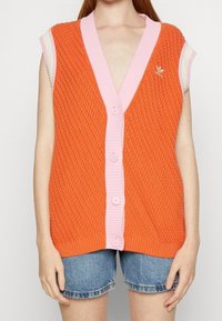 Hihaton oranssi neuleliivi, jossa on vaaleanpunaiset reunat ja napit sekä Adidas-logo, päällä denim-shortsit.