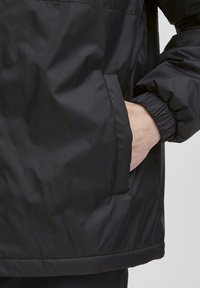 Czarna kurtka przeciwwiatrowa o teksturowanym wykończeniu, z elastycznymi mankietami, bocznymi kieszeniami i prostym dołem, prezentująca minimalistyczny design.