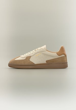 Baskets basses beige et marron clair avec des détails en daim, lacets, perforations et semelle marron, vues de côté sur un fond neutre.