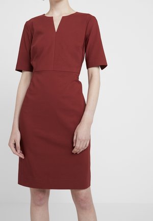 Kvinde iført en knælang, rustfarvet kjole med korte ærmer og en diskret V-udskæring, stående foran en ensartet hvid baggrund.