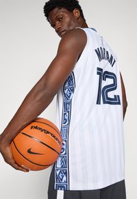 Baltas krepšinio marškinėliai su tamsiai mėlynais akcentais ir geometriniu šonu raštu, su skaičiumi "12" ir "MORANT" ant nugaros. Laiko oranžinį krepšinį.