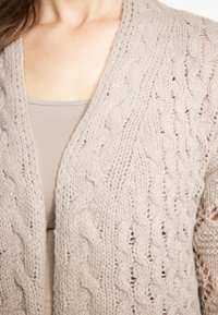 Cardigan lavorato a maglia di un leggero beige con un motivo a treccia, scollatura a V profonda e apertura frontale. Realizzato in un morbido filato testurizzato. Accentuato da un top beige a strati sotto.