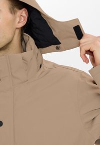 Beige vattentålig jacka med hög krage, tryckknappar och svart vadderad innertyg, designad för skydd mot väder och vind.