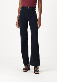 Pantalones de talle alto en color marino con perneras acampanadas, con detalles de botones dorados en los laterales. Llevados con sandalias negras de tacón cuadrado.