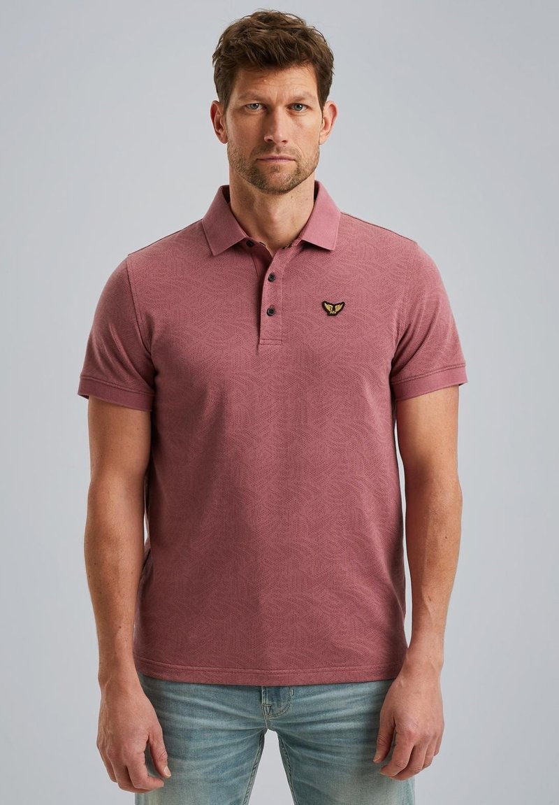 Polo-shirt à manches courtes de couleur rouge clair, avec un motif géométrique texturé, une patte de boutonnage à trois boutons et un petit emblème brodé sur la poitrine.