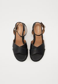Schwarze Sandalen mit Kreuzriemen aus glattem Leder, verstellbarem Knöchelriemen und strukturiertem Schuhboden. Braune Innensohlen mit geprägt eingraviertem Markennamen.