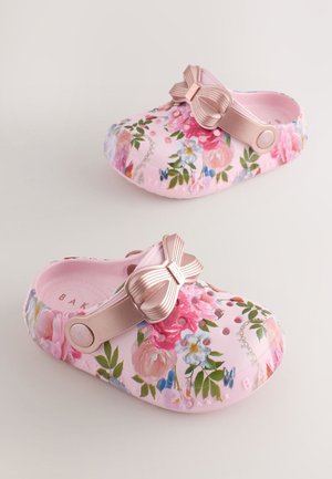 Rosa Blumen-Clogs mit Lüftungslöchern und verstellbaren metallic-pinken Riemen, die dekorative Schleifen auf einem beigen Hintergrund zeigen.